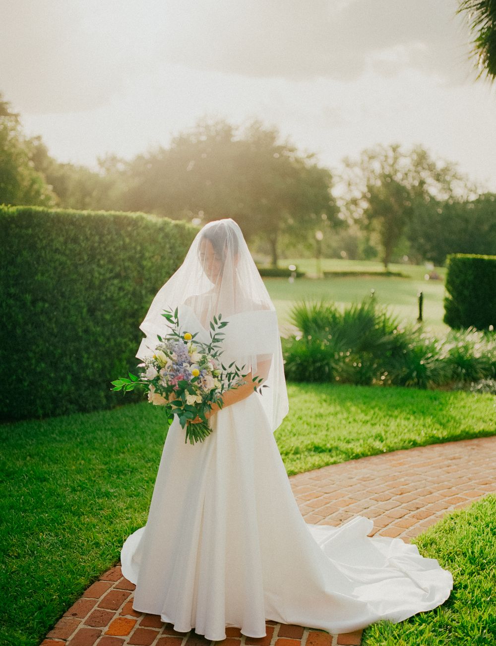 A bride in a flowing white wedding dress and veil walks along a garden pathway on a sunny afternoon.