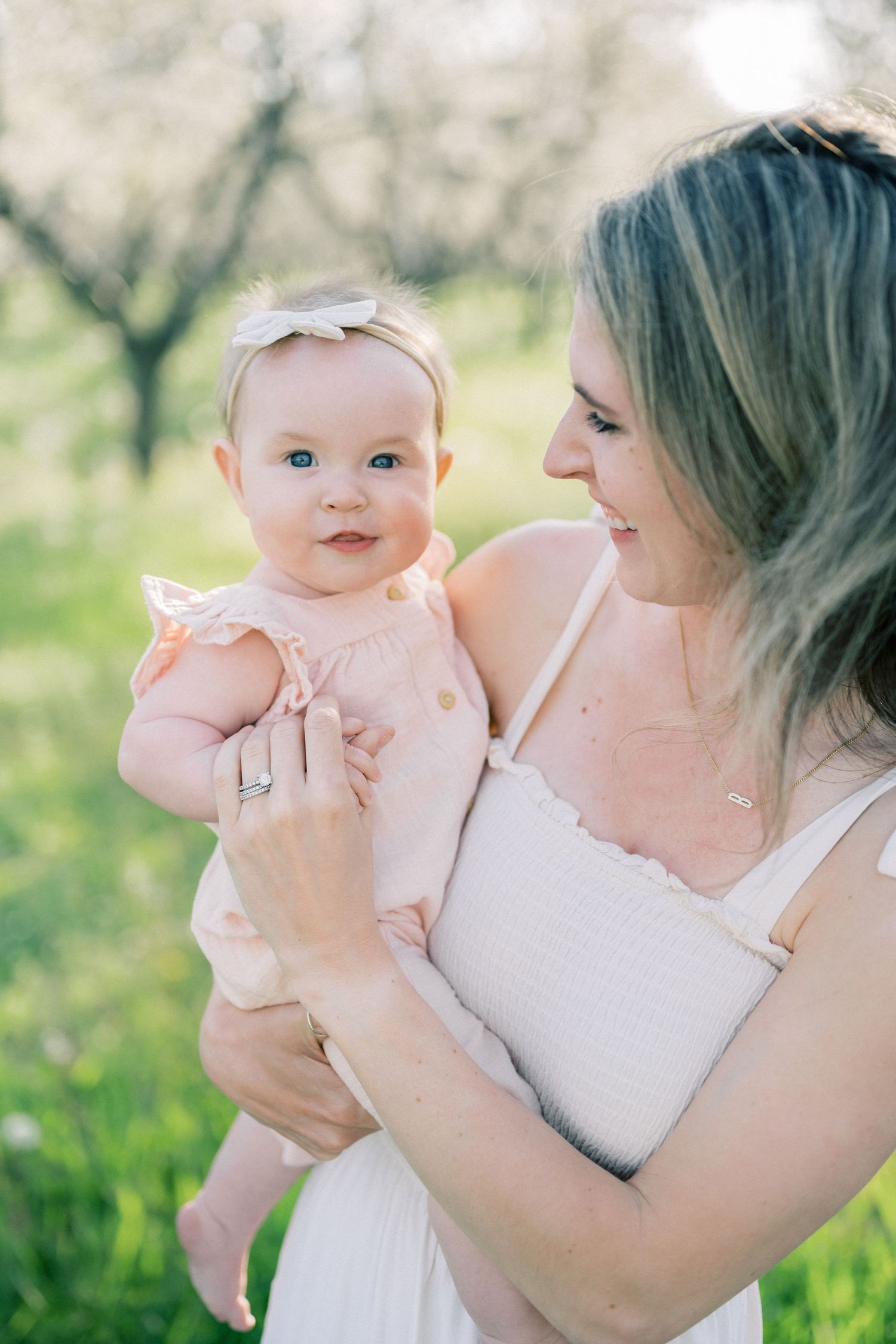 Sweet moment between baby in peach dress with bow headband and parent in white dress standing outdoors in spring garden.