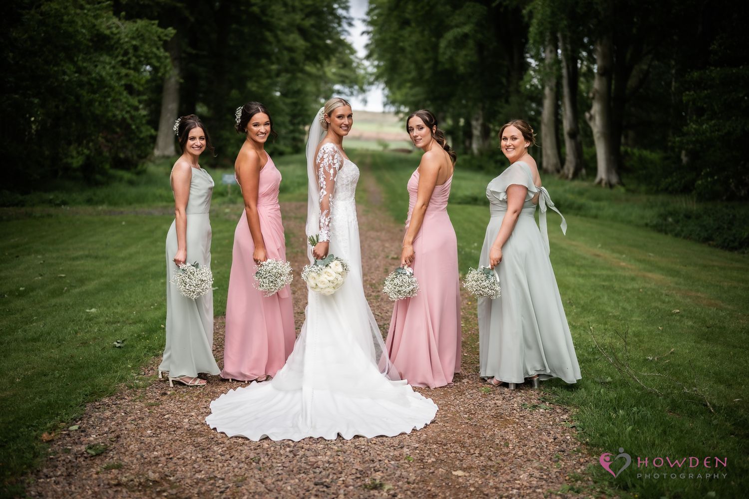 Wedding party in mix of pink and gray bridesmaid dresses standing on garden path surrounded by trees.