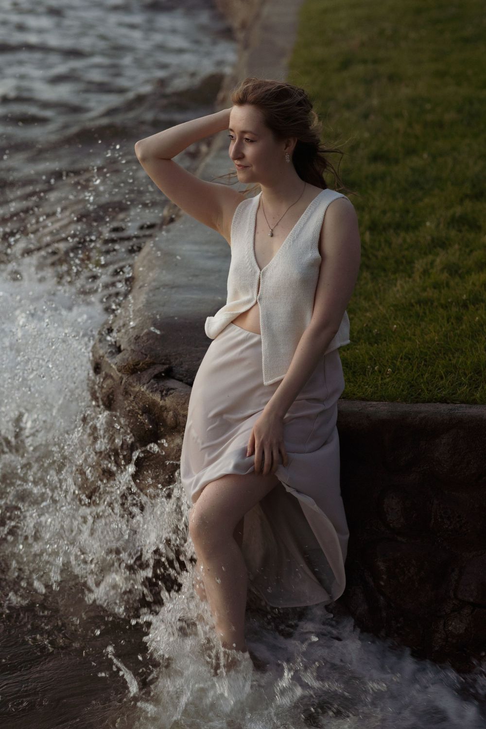 Woman in light dress poses near crashing waves along shoreline.