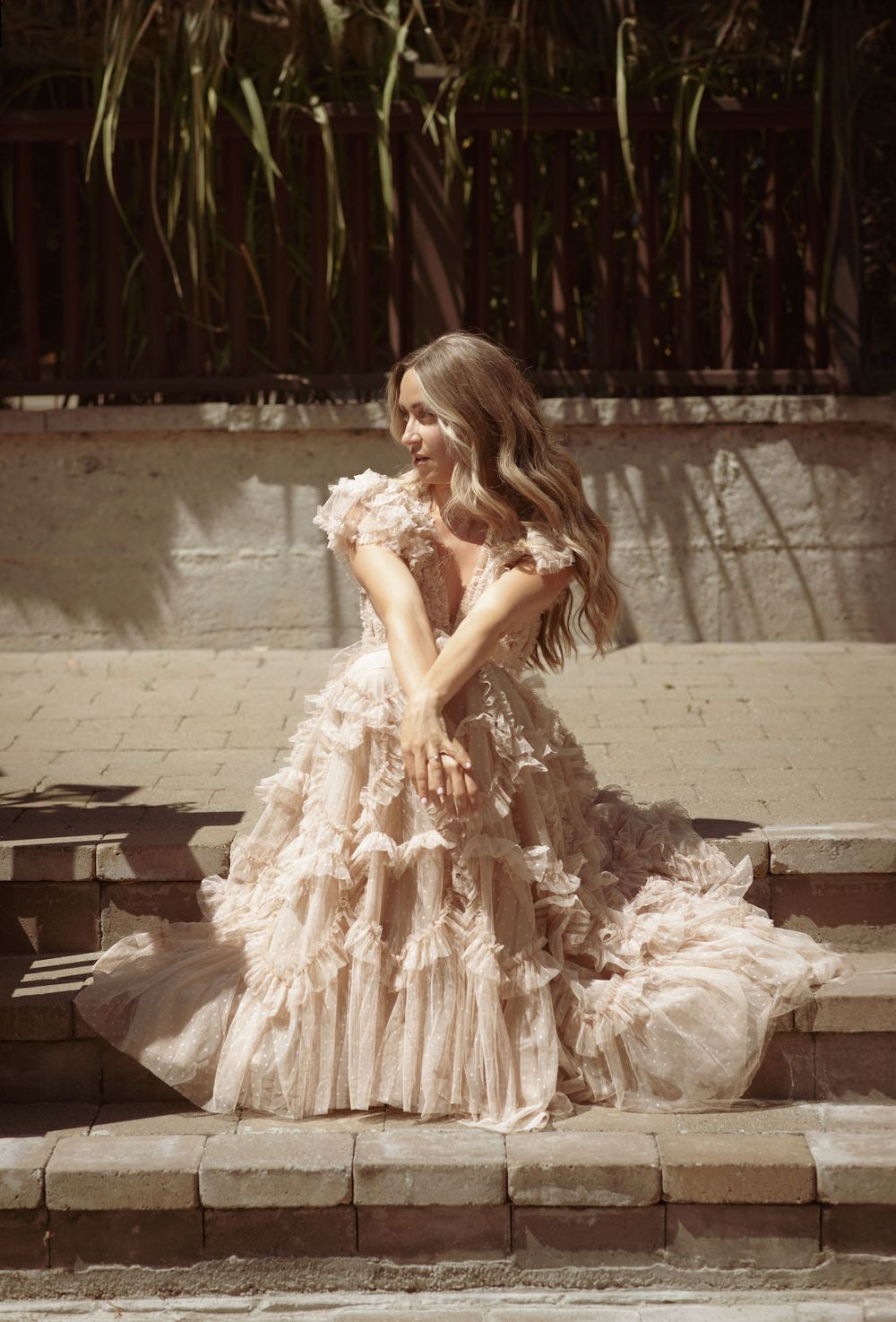 Elegant ruffled blush gown catches sunlight against textured stone wall backdrop.