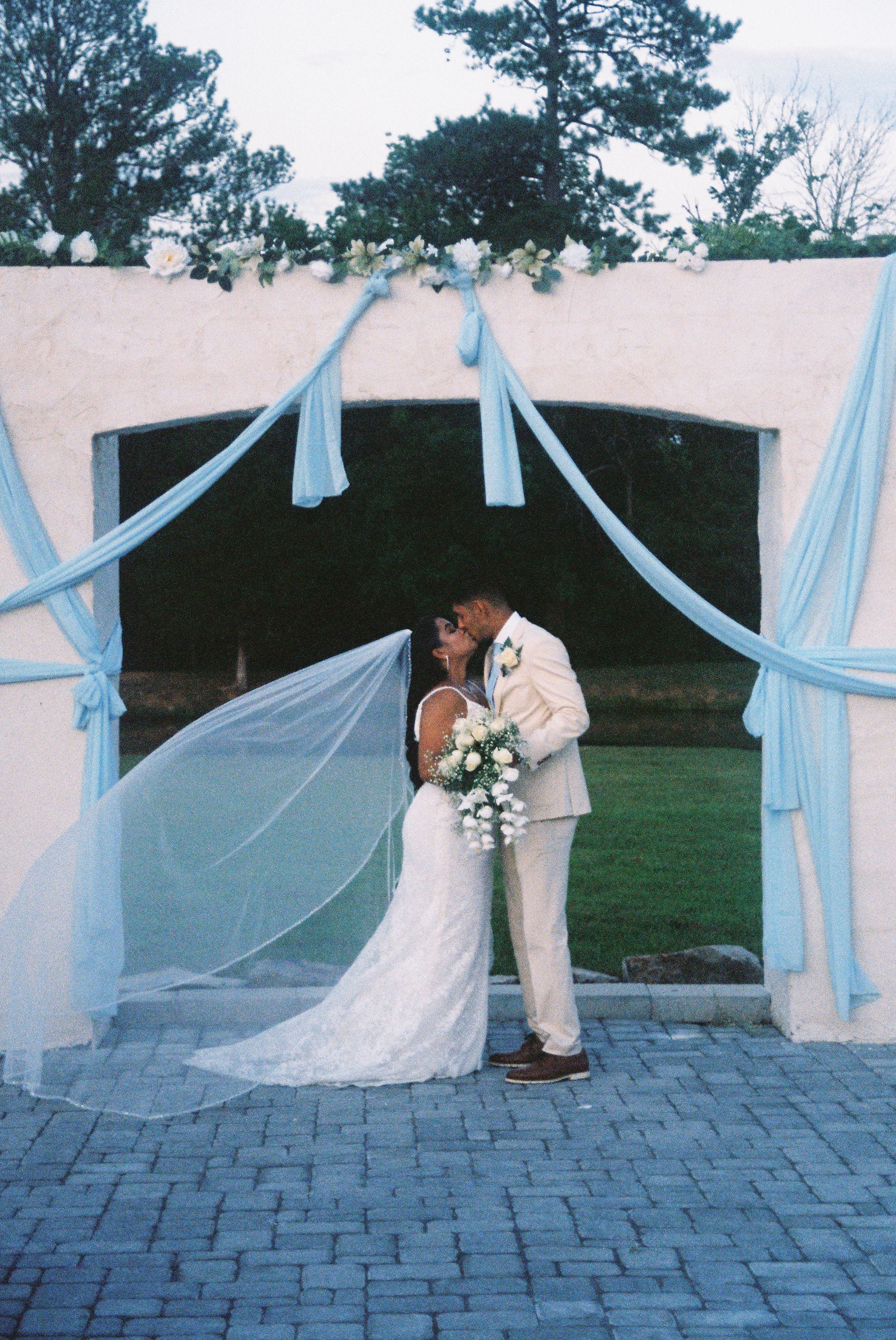 A newlywed couple shares a romantic moment under light blue drapery at their outdoor wedding ceremony setup.