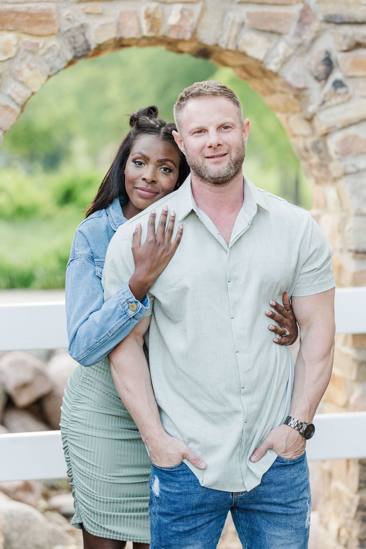 A couple poses together outdoors in front of a stone archway while wearing casual clothing.