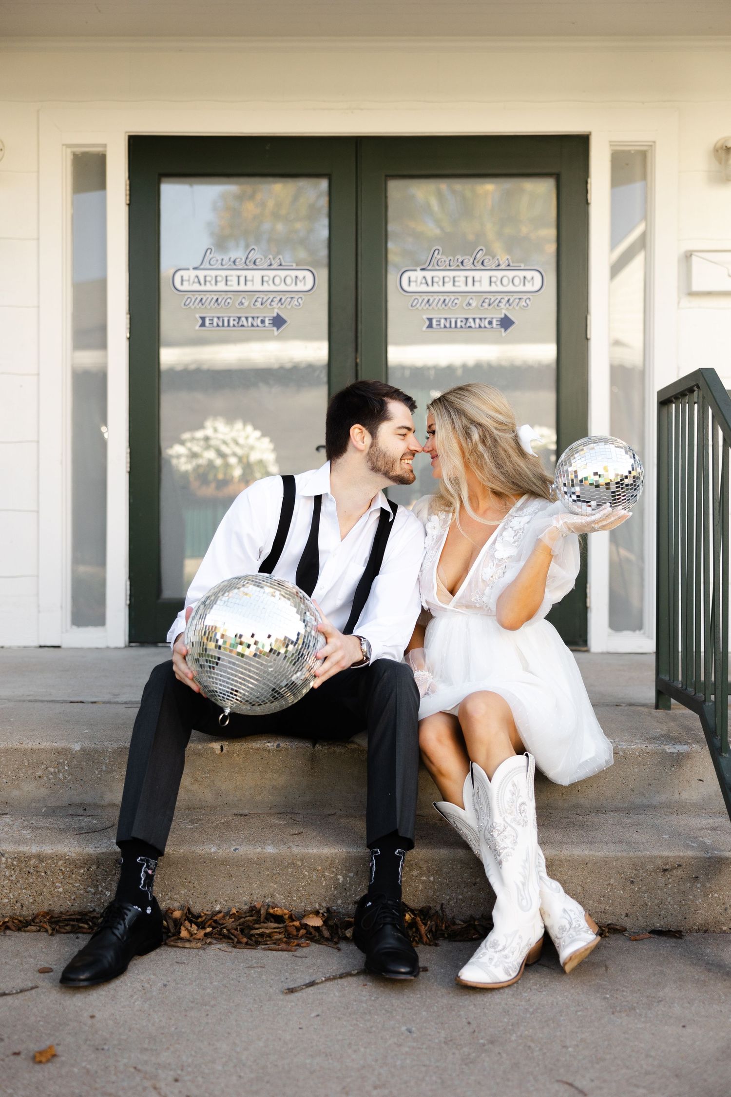 Two people sit on steps holding disco balls while dressed in white and black attire outside a storefront.