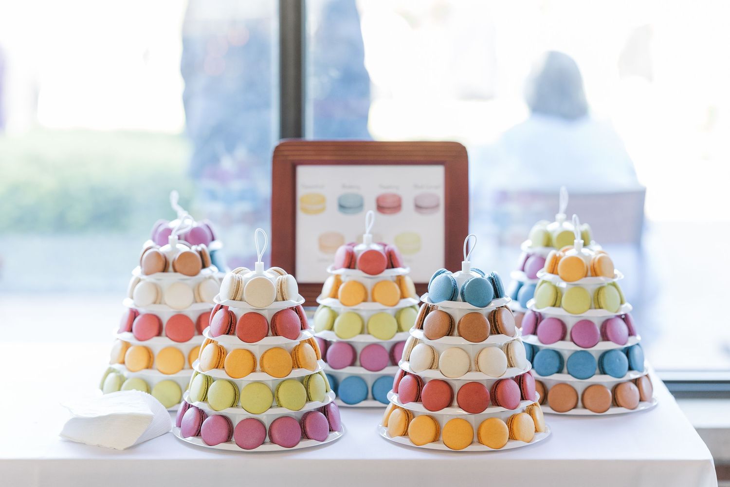 Colorful macarons arranged in pyramid displays on a white table by a window create an elegant pastel dessert presentation.
