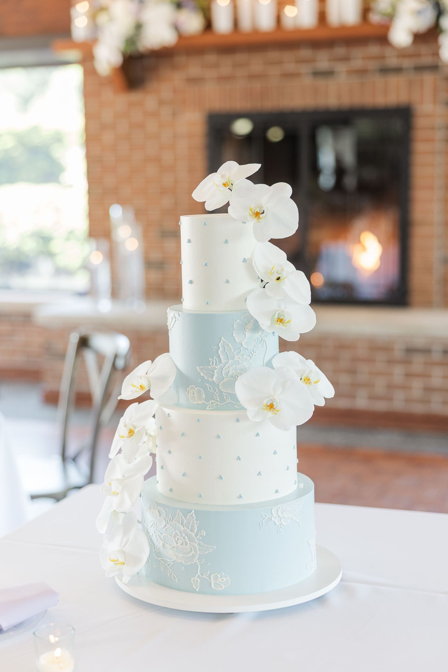A light blue and white wedding cake decorated with delicate lace patterns and fresh white orchids on a silver cake stand.