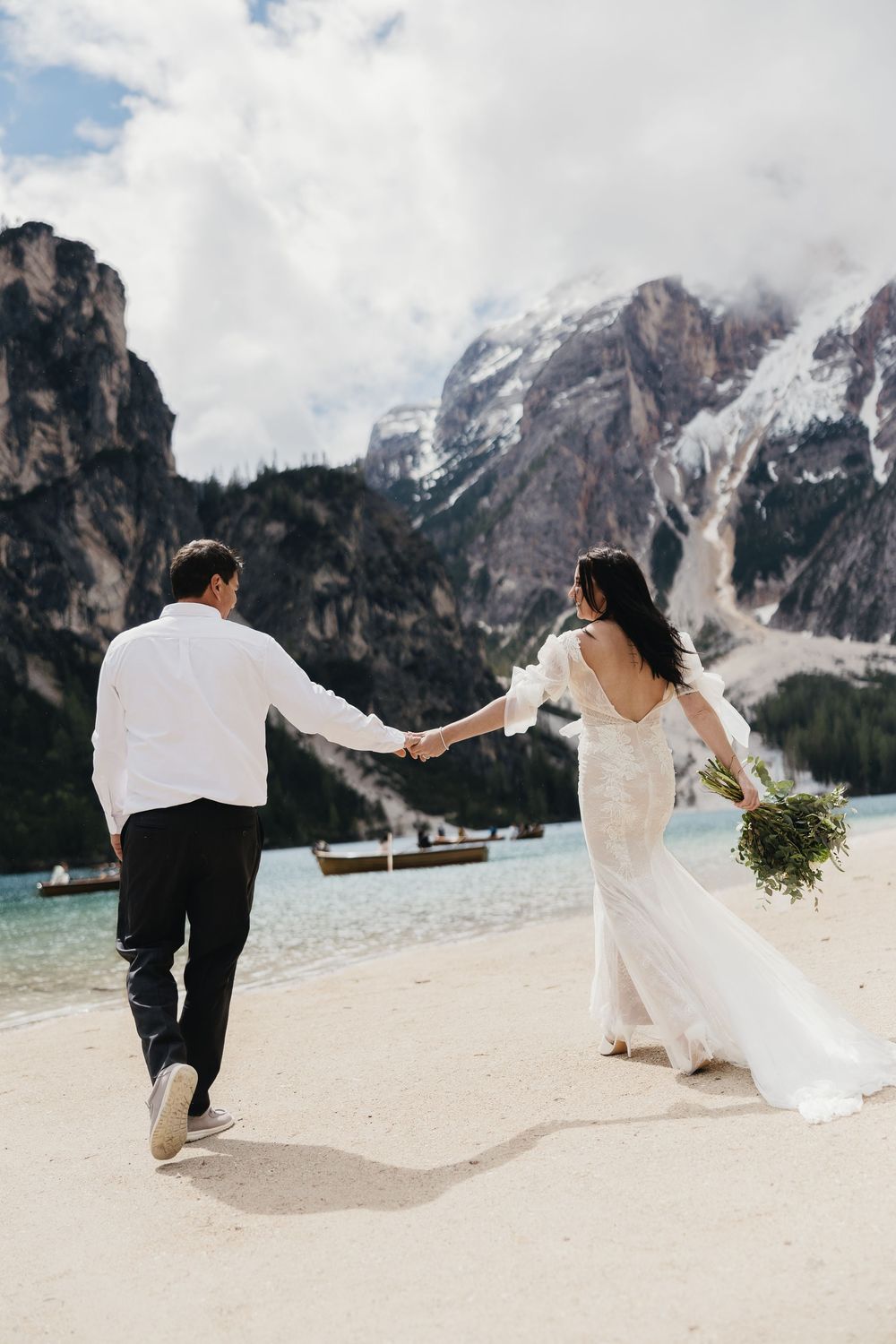 Newlyweds holding hands while running on a beach with dramatic mountain peaks and turquoise lake in the Dolomites.