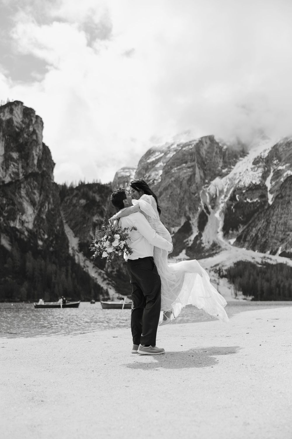 A romantic couple embraces against a dramatic mountain backdrop with a scenic lake and cloudy sky at Lago di Braies.