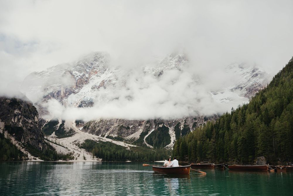 Wooden boats float on a misty alpine lake surrounded by snow-capped mountains and evergreen forest in the Dolomites.
