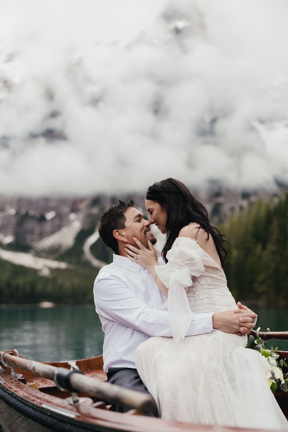 A romantic couple shares tender moments in a boat on a misty mountain lake with evergreen trees in the background.