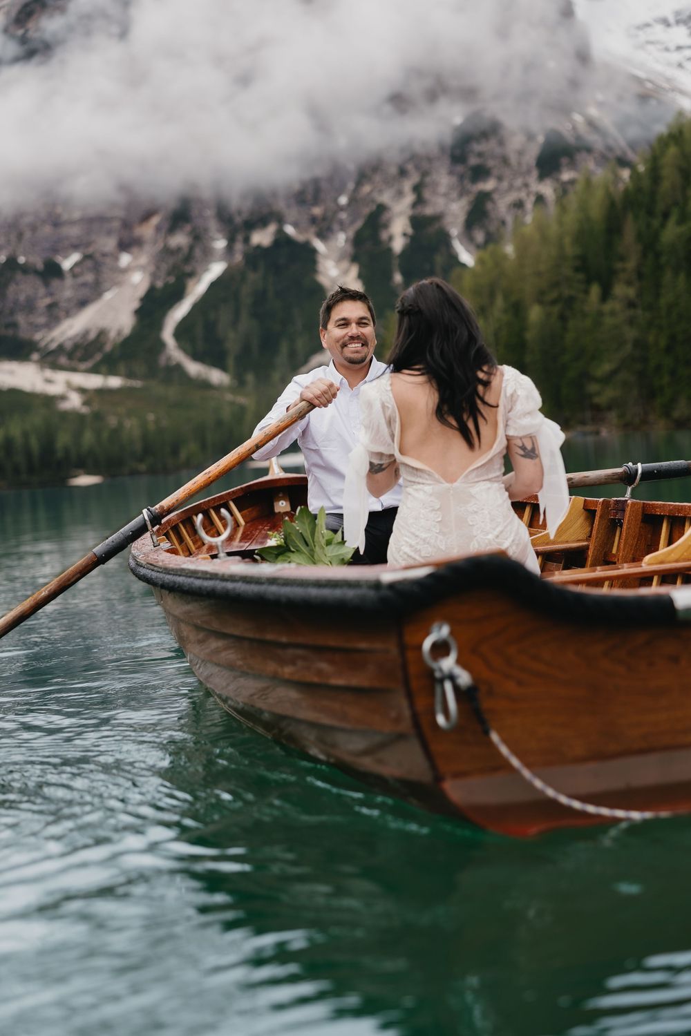Couple sharing romantic moment in wooden rowboat on turquoise alpine lake surrounded by misty Dolomite mountains.