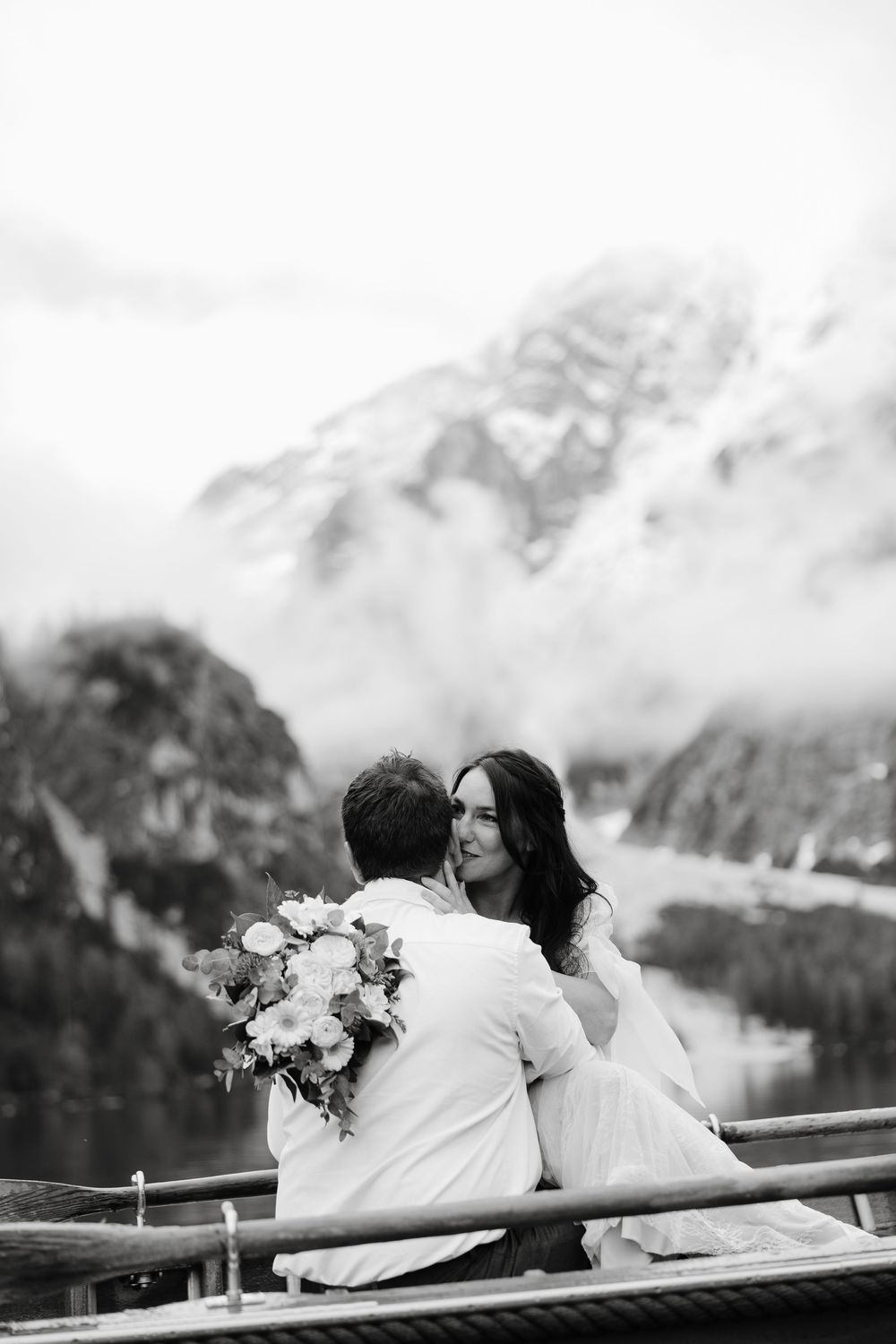Black and white photo of a couple sitting on a dock overlooking misty mountain scenery.