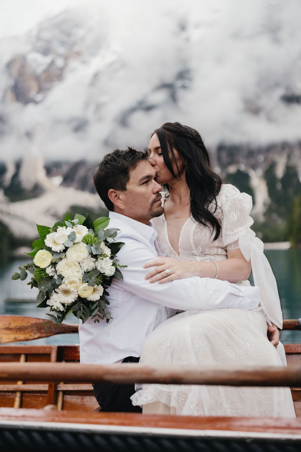 Newlyweds share an intimate moment on a wooden boat with snow-capped mountains and a white floral bouquet in view.