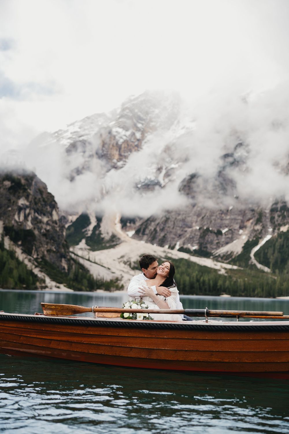 Wedding couple share an intimate moment in wooden boat on alpine lake surrounded by misty mountain peaks.