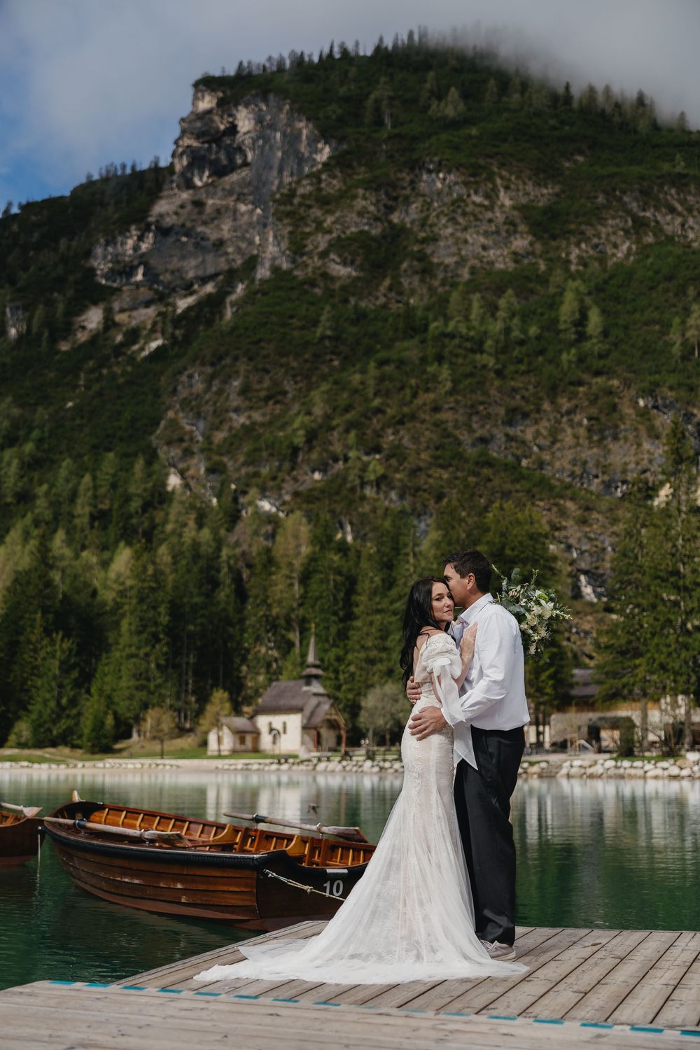 A couple in wedding attire embraces on a wooden dock beside a lake surrounded by majestic mountain cliffs and pine trees.
