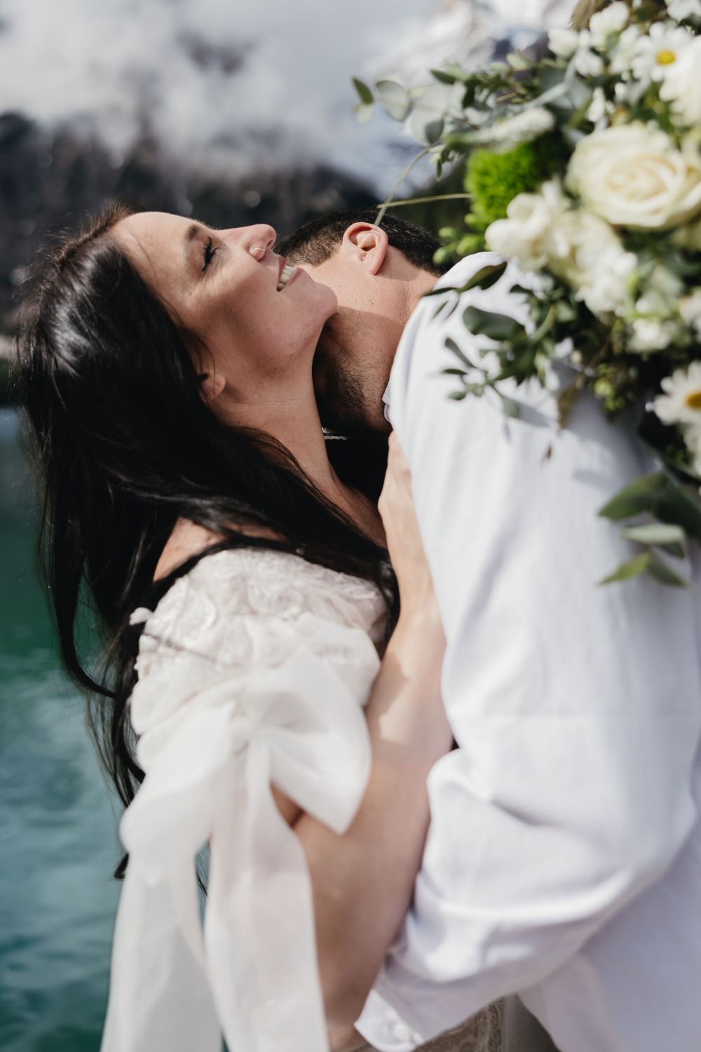 Newlyweds share intimate moments surrounded by white flowers at a lakeside wedding ceremony.