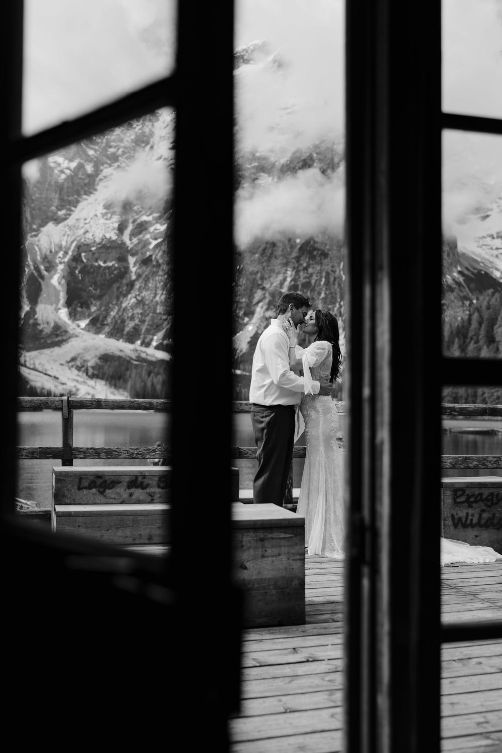 Black and white photo of a couple embracing on a deck overlooking majestic mountain peaks and a lake.