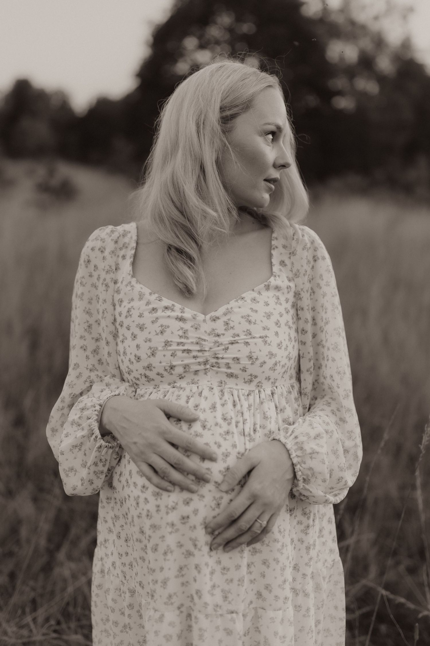 Black and white portrait of a blonde woman in a floral dress standing in a field looking to the side.