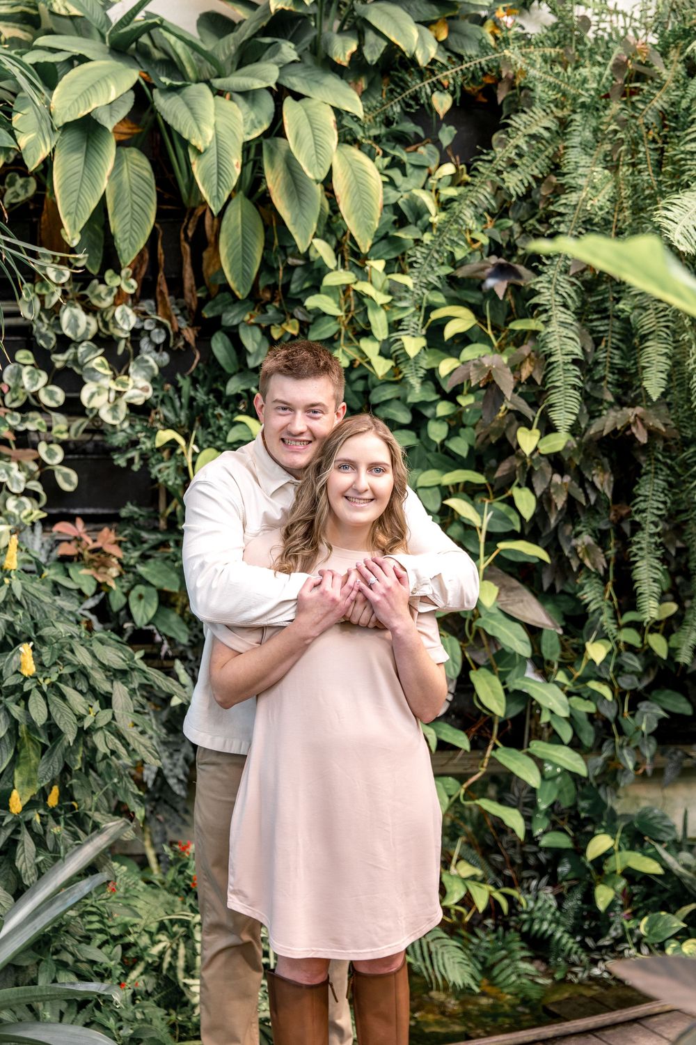 A couple embraces in front of a lush green wall of tropical plants and foliage in a greenhouse setting.