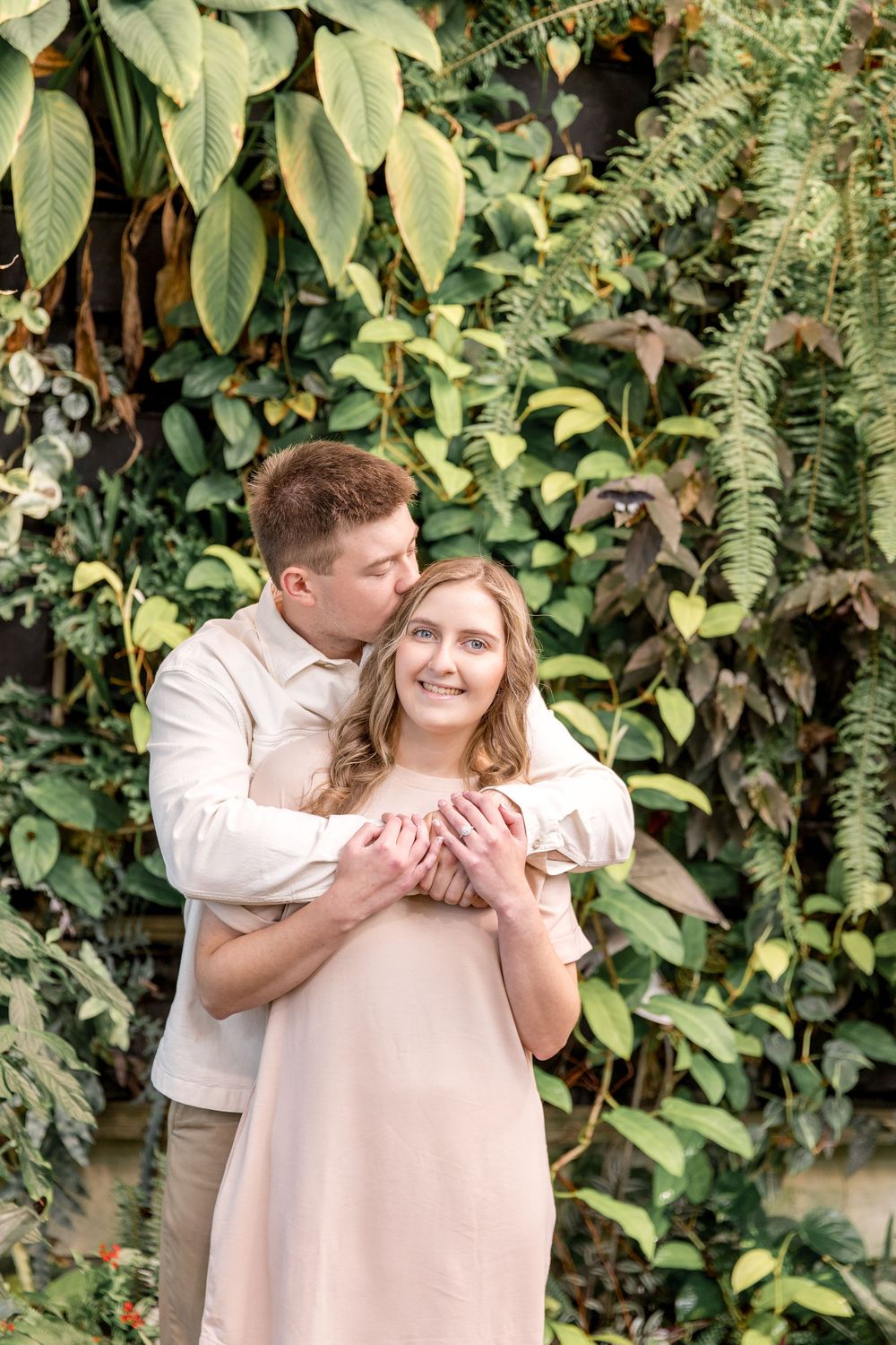 A loving couple embraces in front of a lush green wall of tropical plants and foliage.