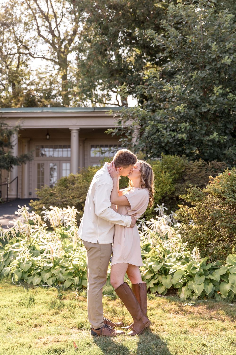 A romantic couple shares a tender moment in a sunlit garden with a beautiful columned building in the background.