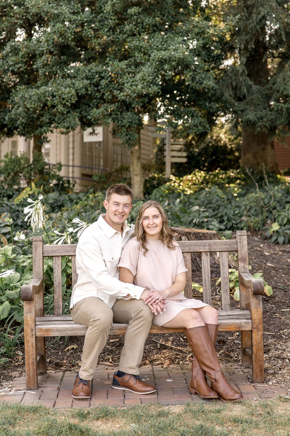A couple sits together on a wooden garden bench surrounded by lush greenery and trees in a romantic outdoor setting.