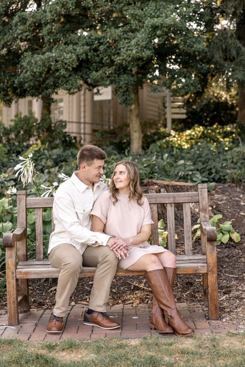 A couple sits together on a wooden garden bench surrounded by lush greenery and trees in a romantic outdoor setting.