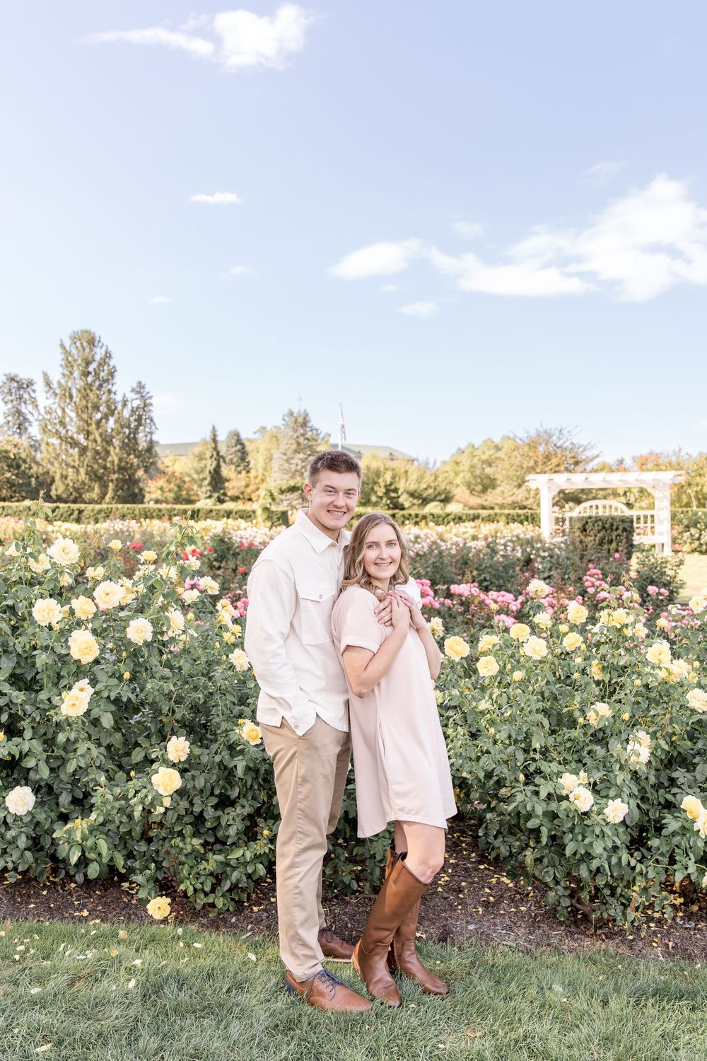 A couple poses together in a beautiful rose garden with blooming yellow flowers on a sunny afternoon.