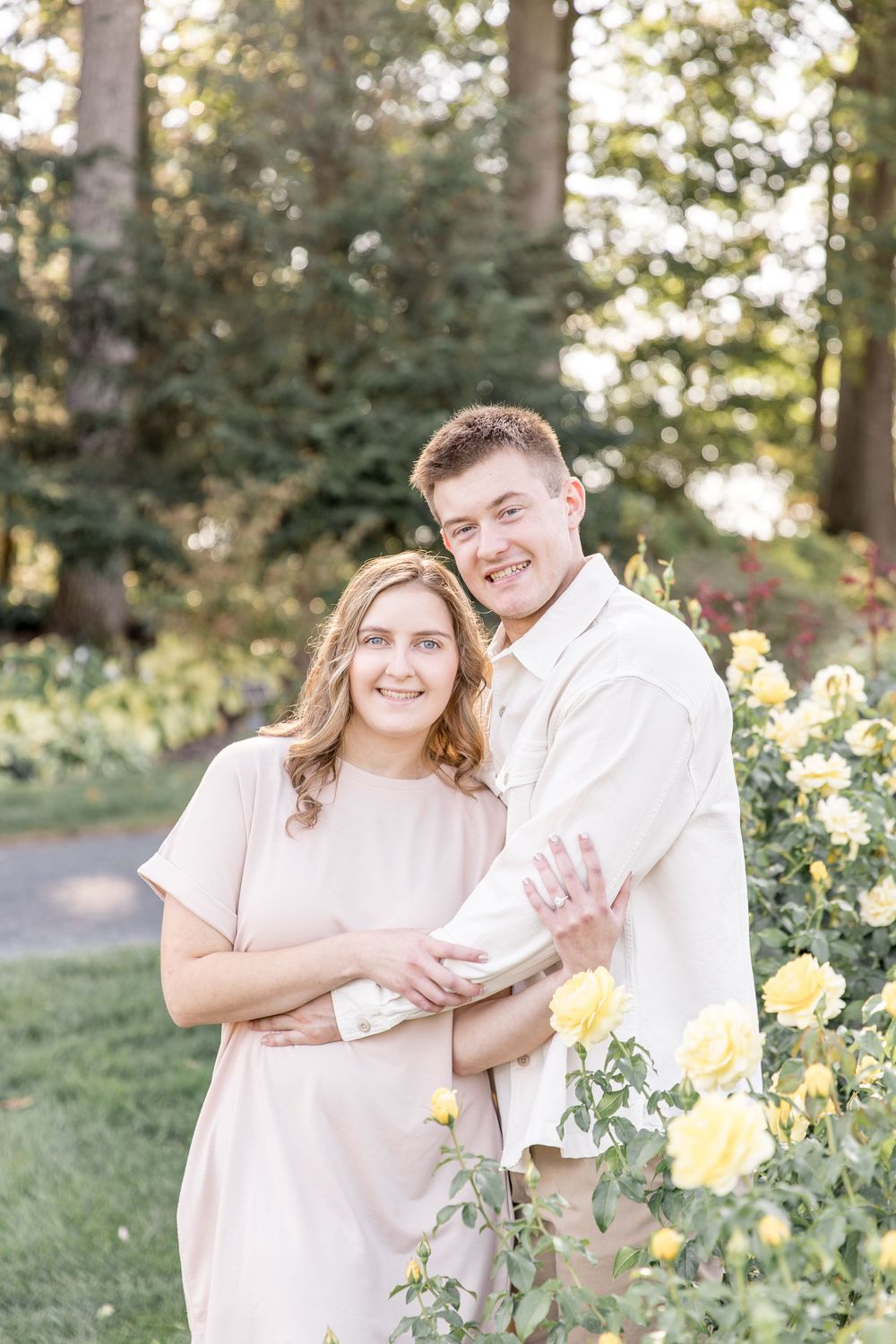 A couple embraces among yellow flowers in a garden setting with soft natural lighting and neutral clothing.