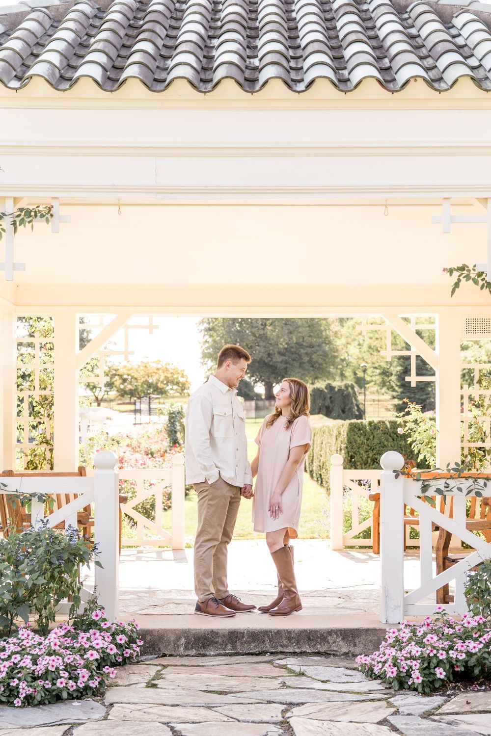 A couple stands facing each other in a garden gazebo surrounded by pink flowers and Spanish tile roofing.