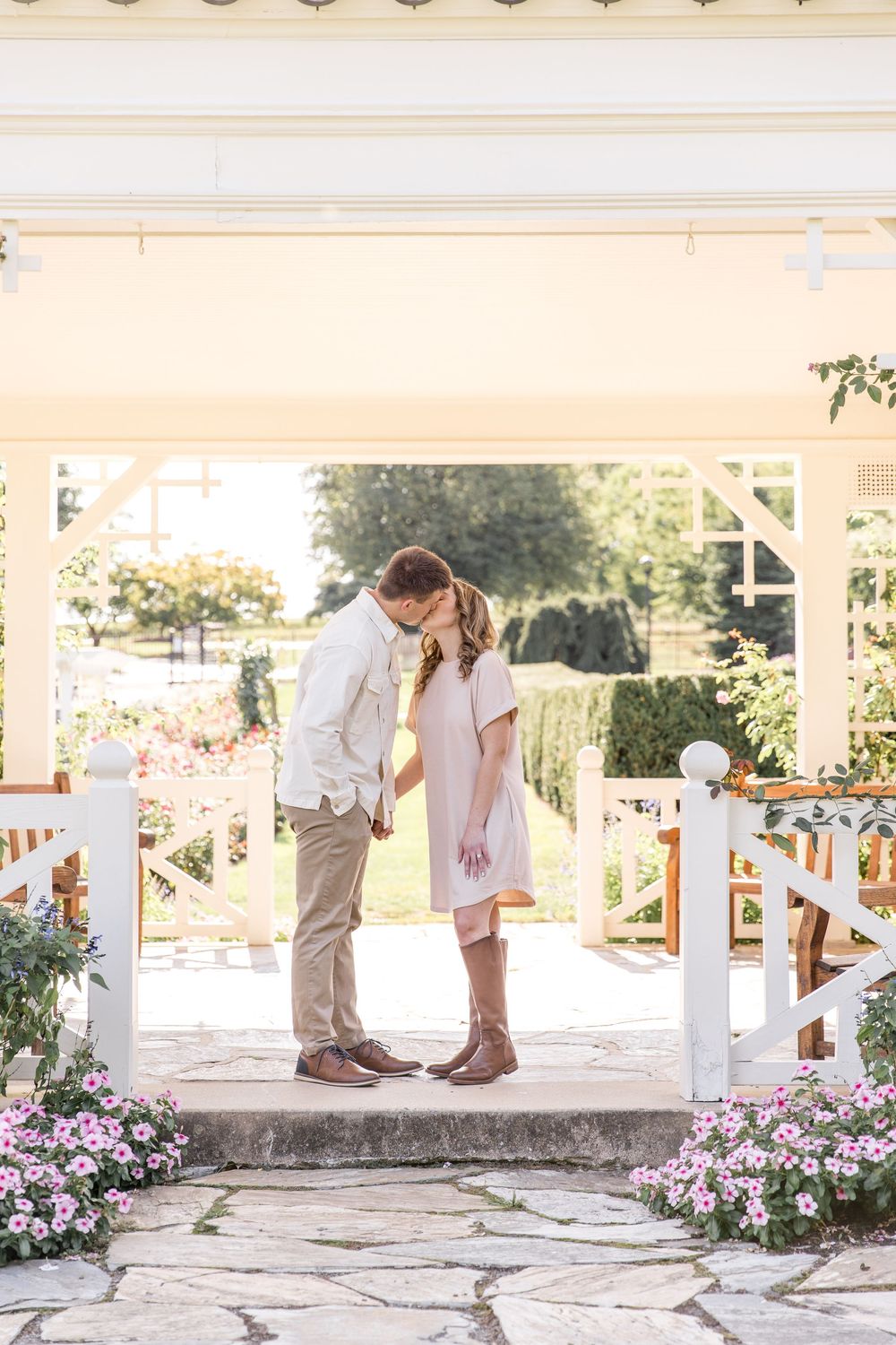 A romantic couple shares an intimate moment on a stone patio surrounded by pink flowers and a white pergola.