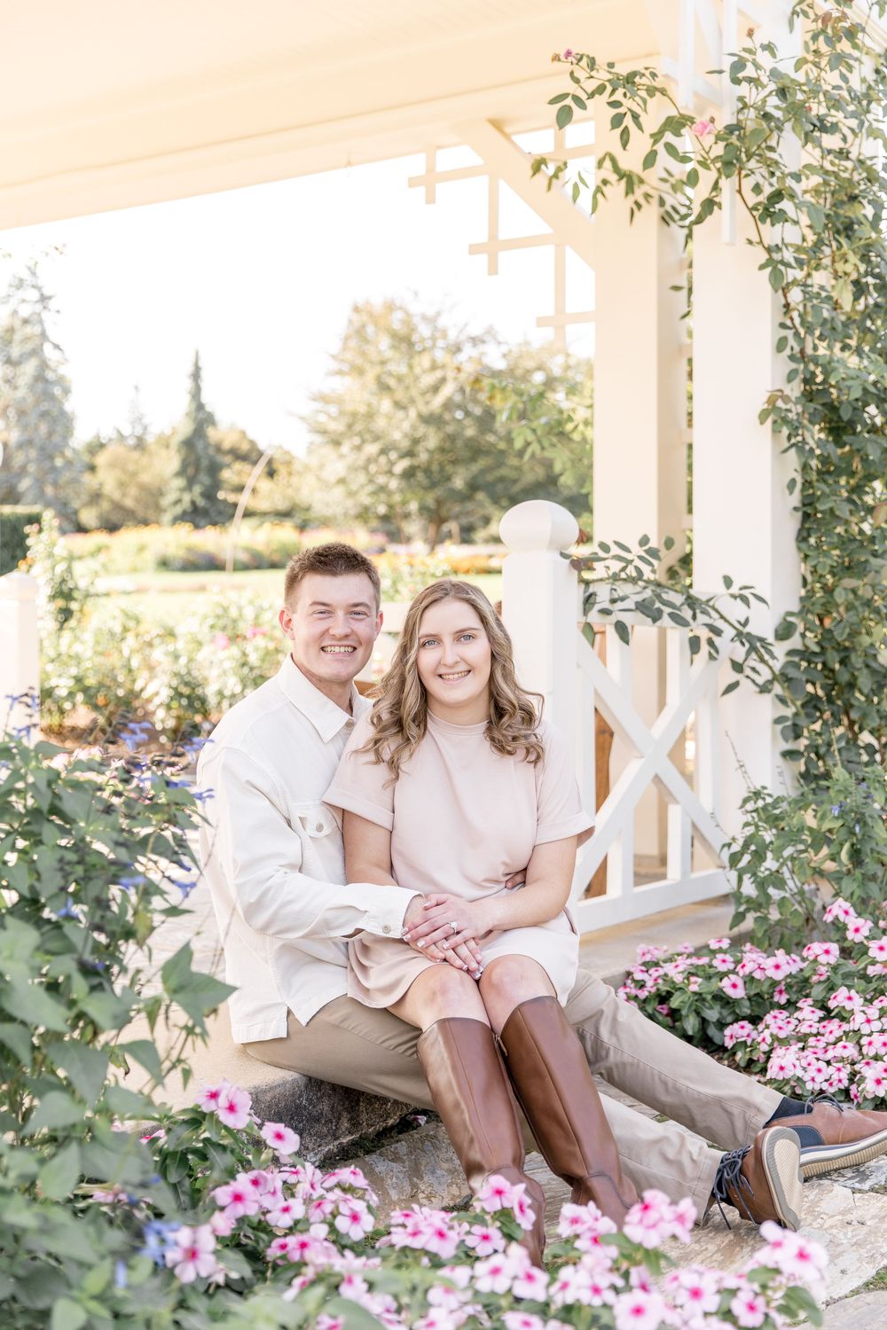 A happy couple sits on garden steps surrounded by pink flowers and greenery on a sunny day.