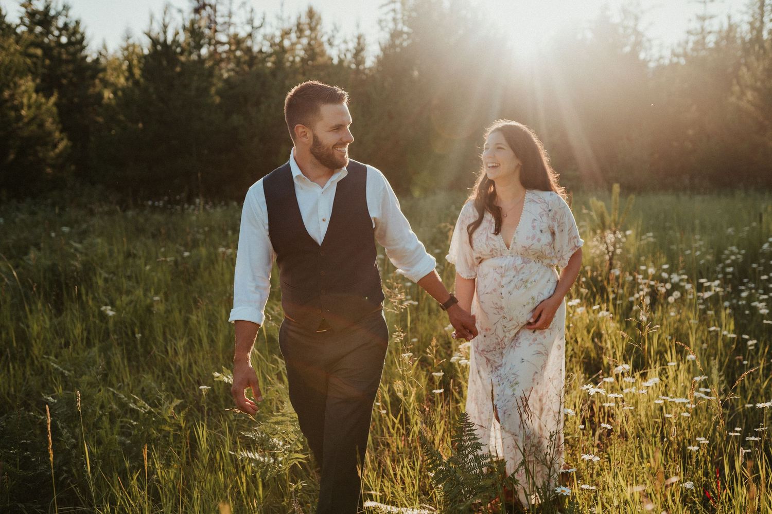 Couple walking hand in hand through sunlit meadow during golden hour in rustic wedding attire.
