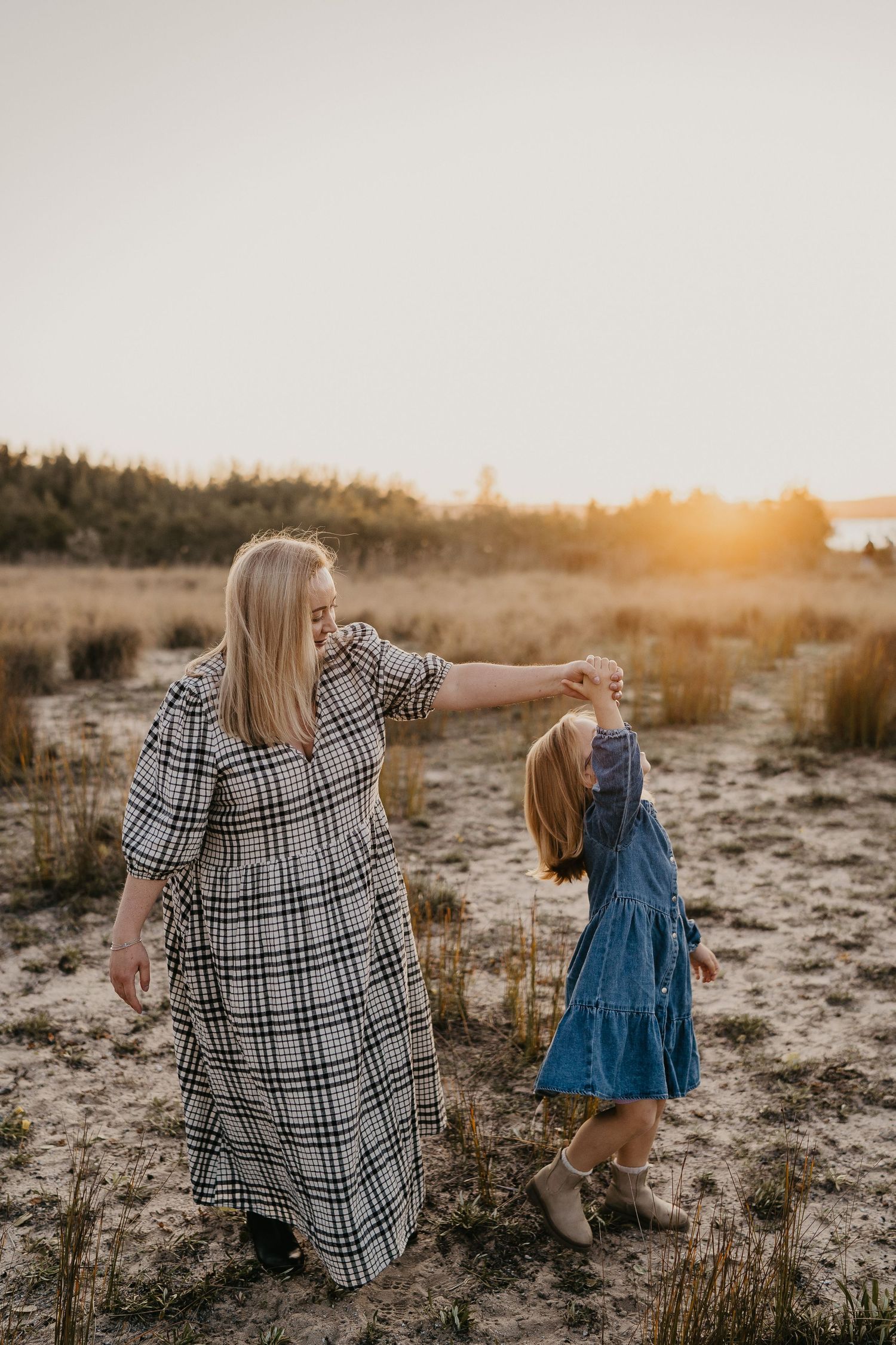 Mother in plaid dress and child in blue dress holding hands while walking in a sandy field at golden sunset.