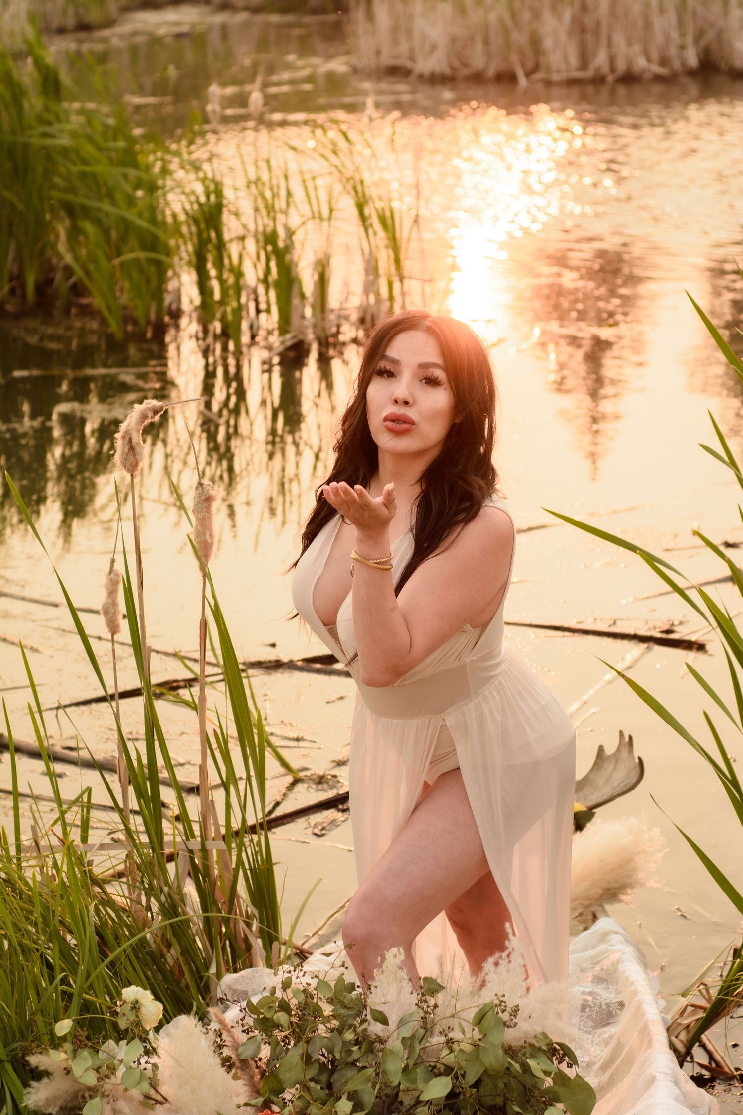 A romantic portrait by a pond with sunset light shining through reeds and wildflowers in a dreamy summer setting.