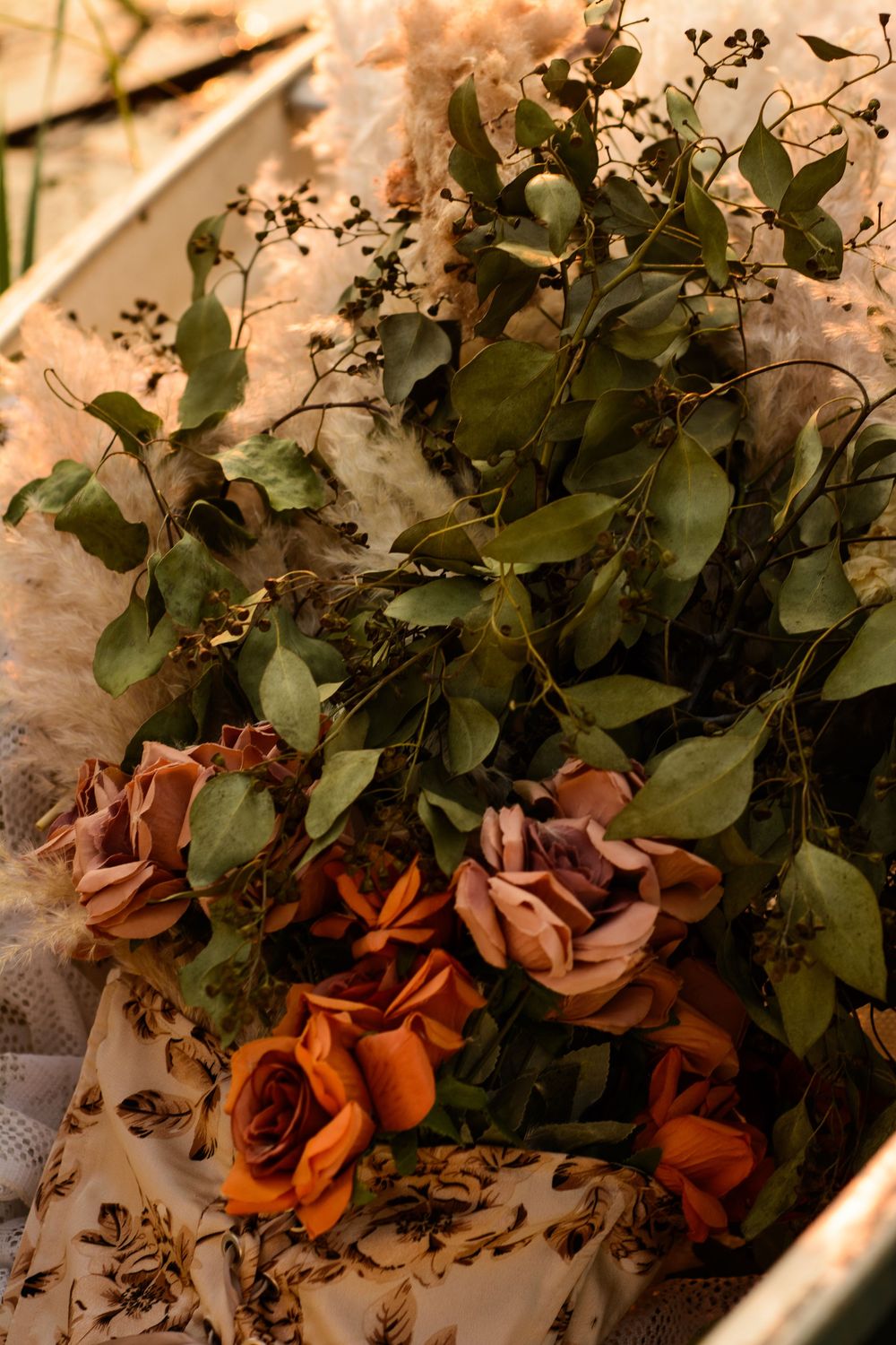 Dried brown roses and green foliage arranged in a decorative display against a cream colored background.