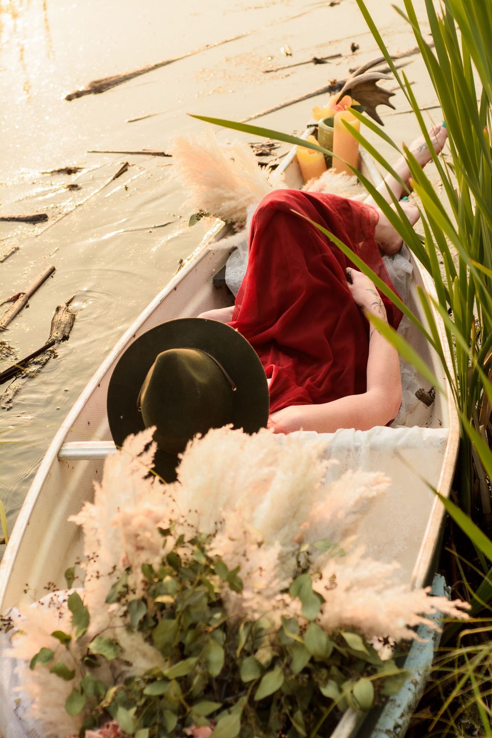 A person in a red dress lying in a boat with dried flowers and reeds along a riverbank.