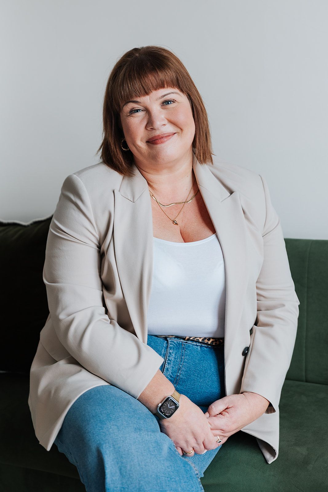 A person in a beige blazer and jeans sits smiling on a green couch against a grey background.