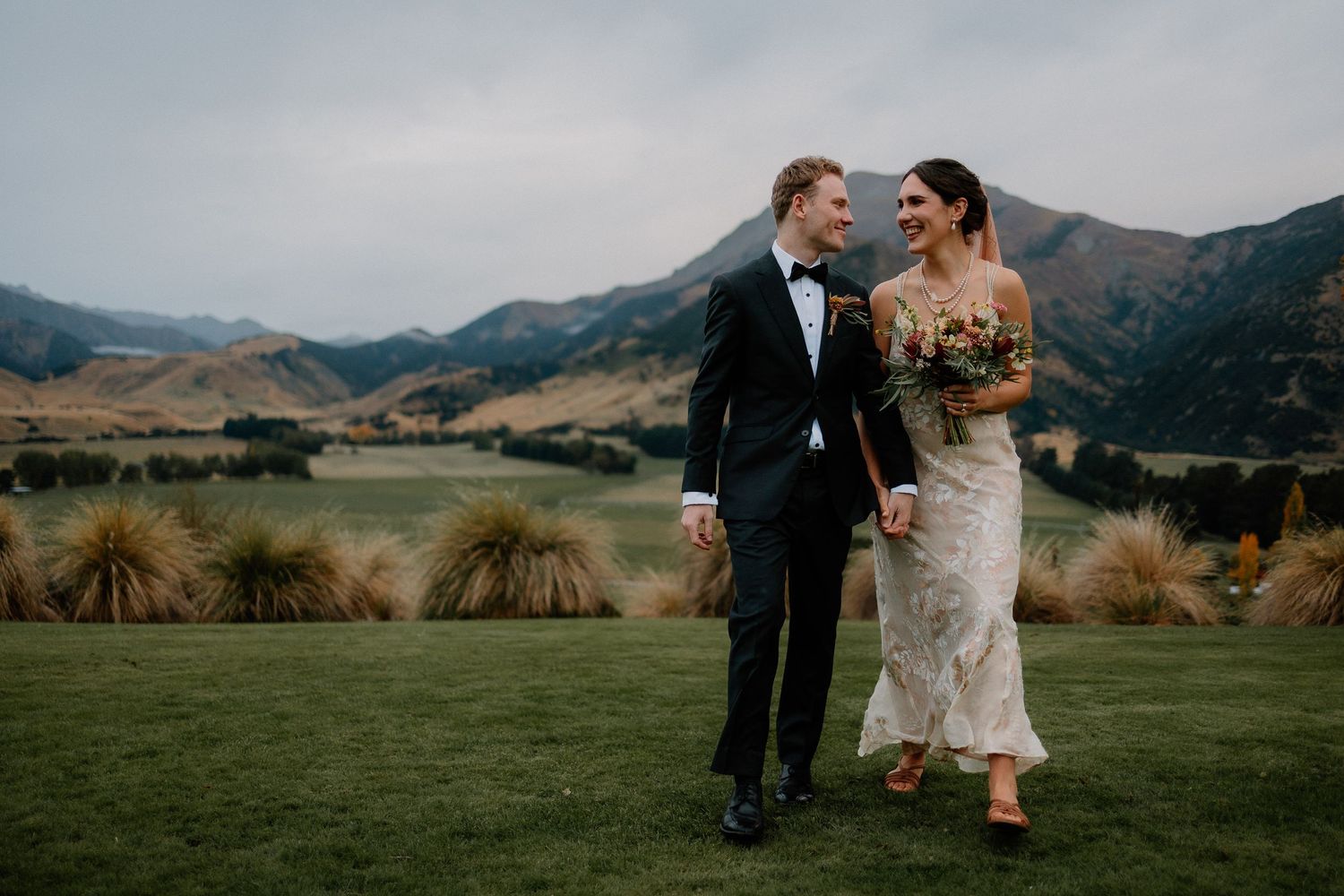 Newlywed couple walks through scenic mountain meadow at sunset with dramatic landscapes in the background.