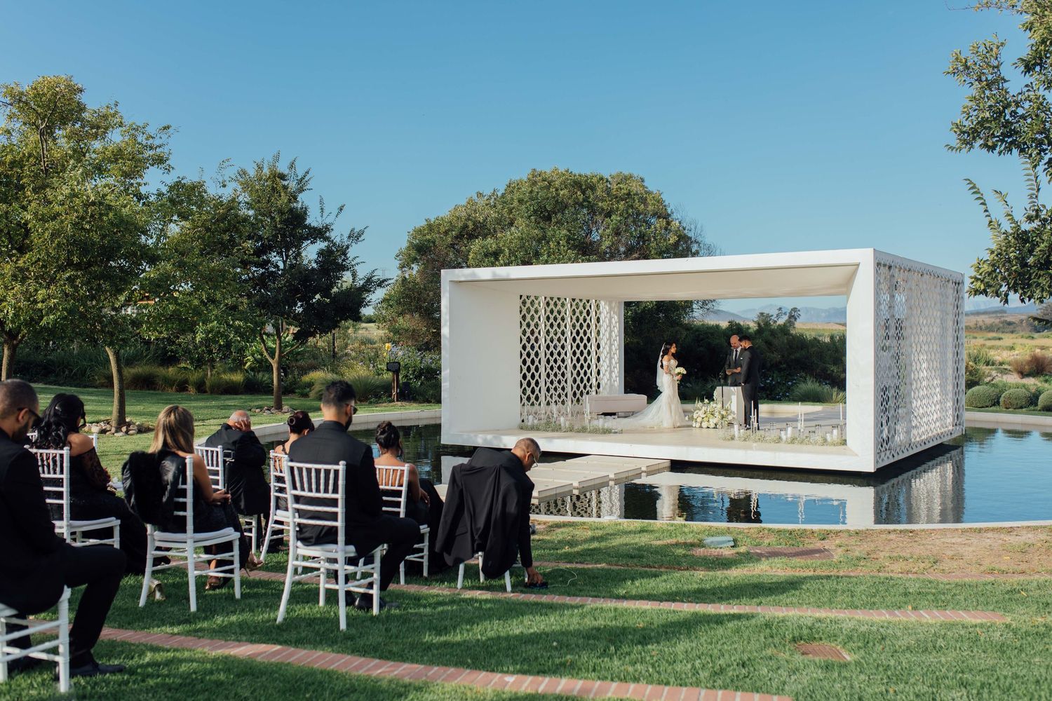 A modern white wedding pavilion overlooks a pond with white chairs set up for an outdoor ceremony.