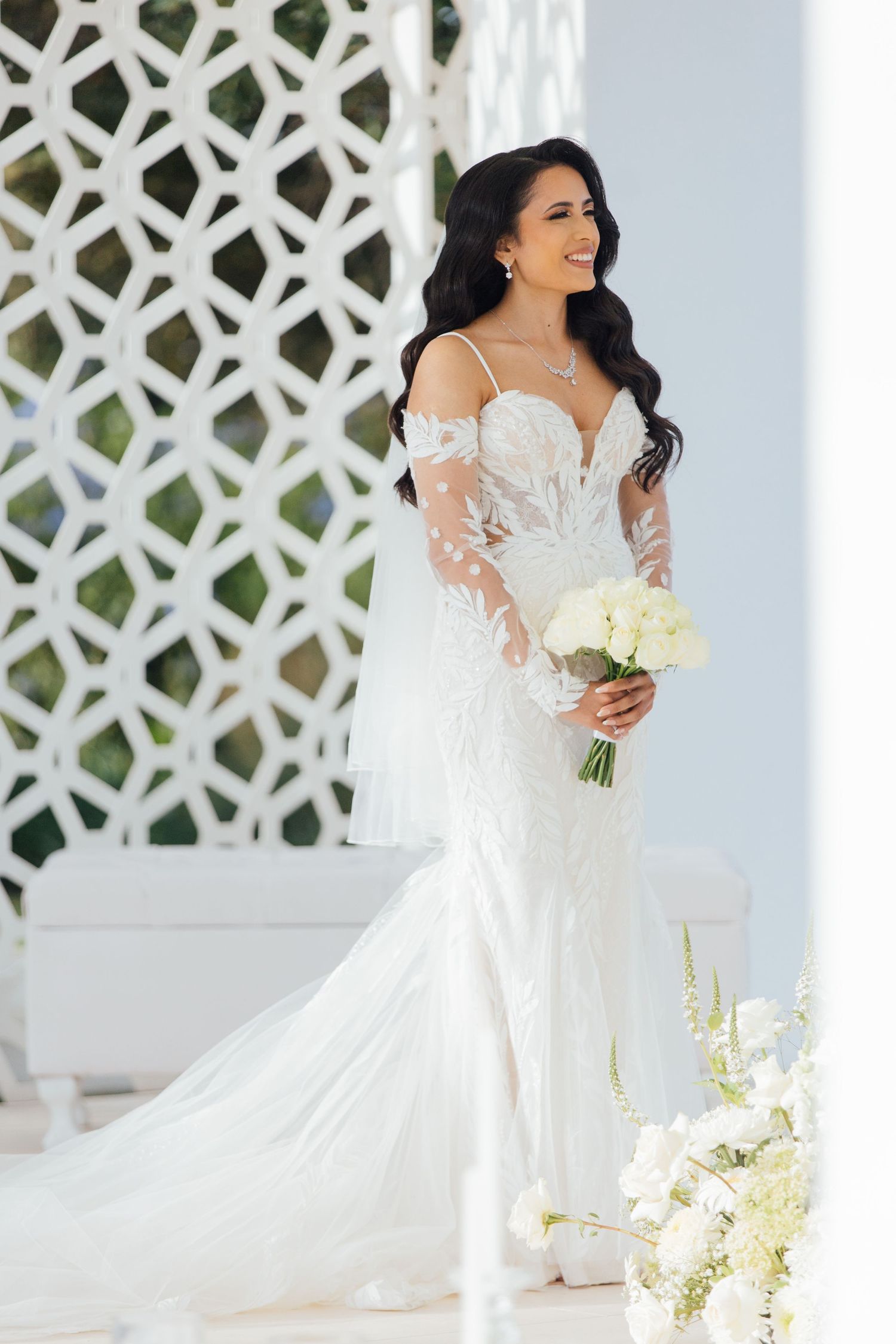 A bride in a fitted lace wedding gown with sheer long sleeves holds a white bouquet against a decorative lattice wall.