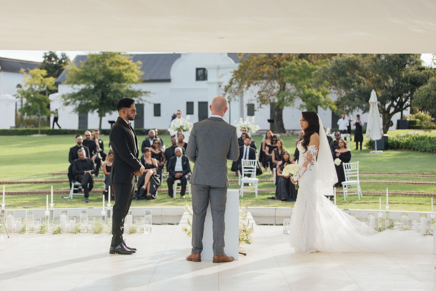 An outdoor wedding ceremony taking place on a white platform with chairs set up on a lawn in front of a white building at Hazendal.