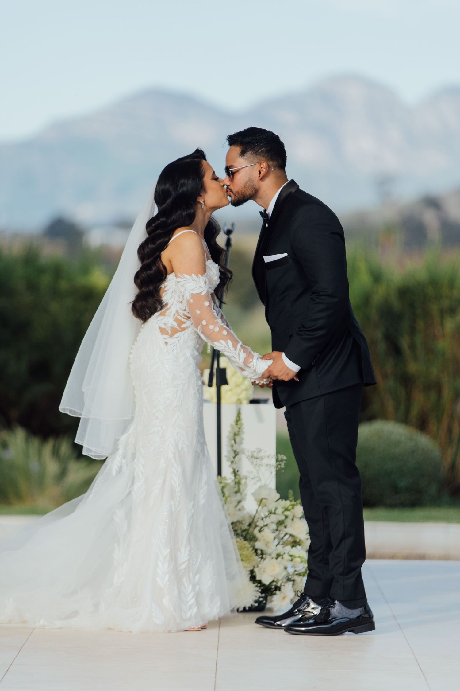 A couple shares their first kiss as newlyweds on an outdoor terrace with mountains in the background.