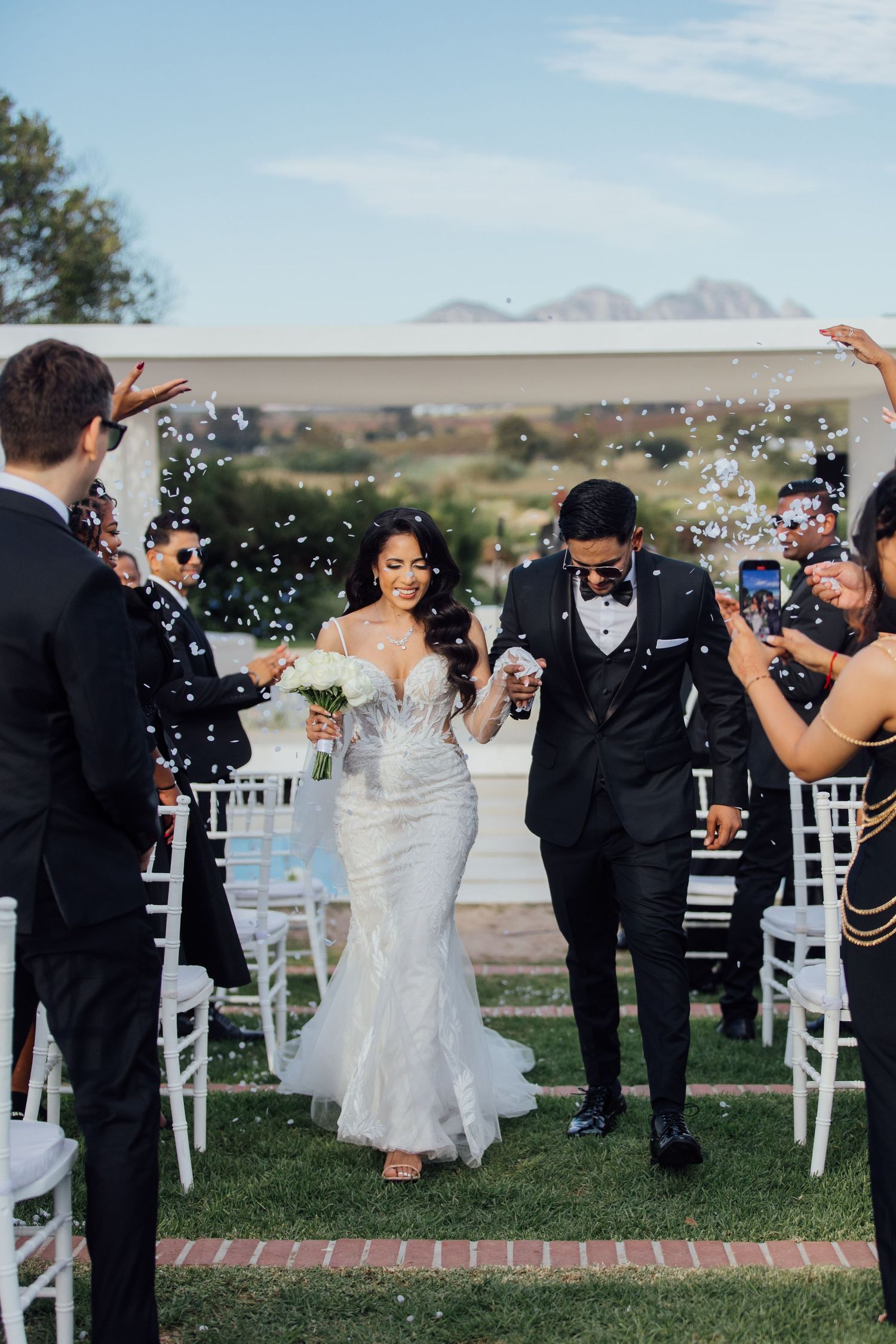 Newlyweds walk down aisle at outdoor wedding ceremony with mountains in background as guests shower them with confetti.