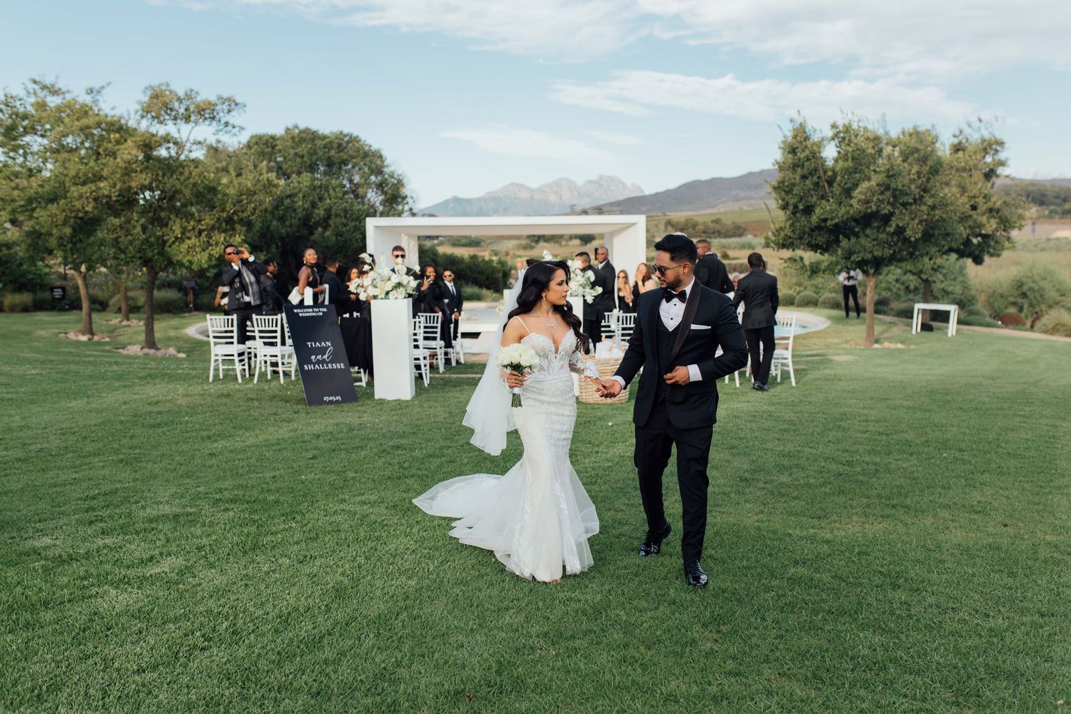A couple walks joyfully across a lush green lawn after their outdoor wedding ceremony with guests and decor in background.