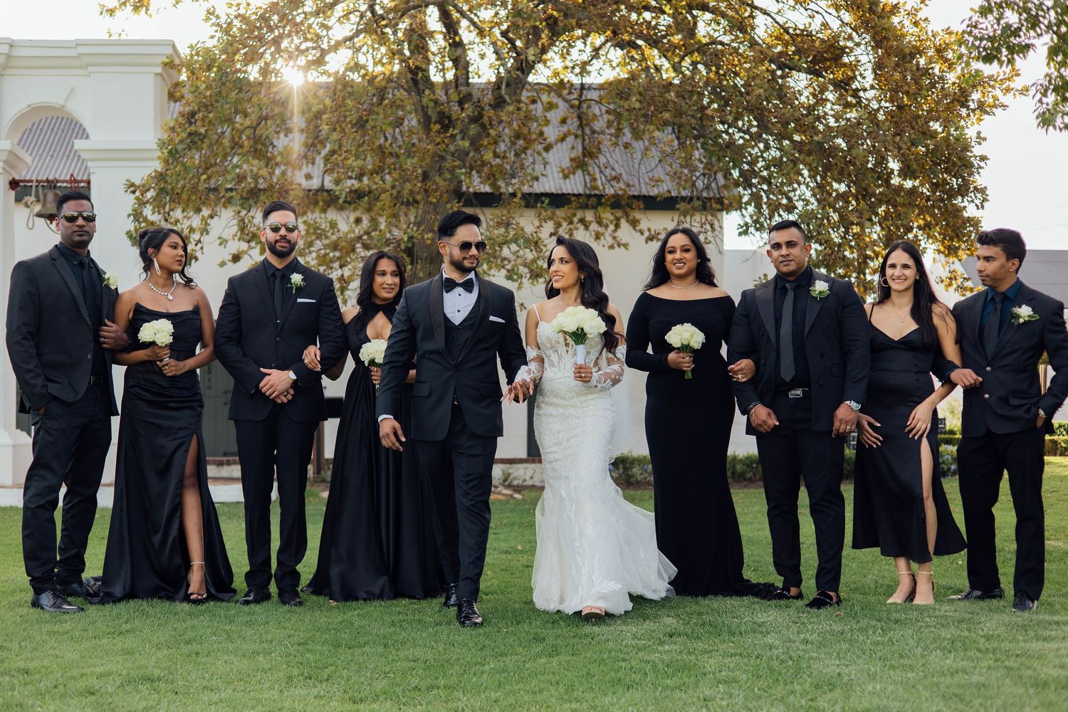 A wedding party wearing black and white formal attire gathers on a green lawn during an outdoor ceremony.