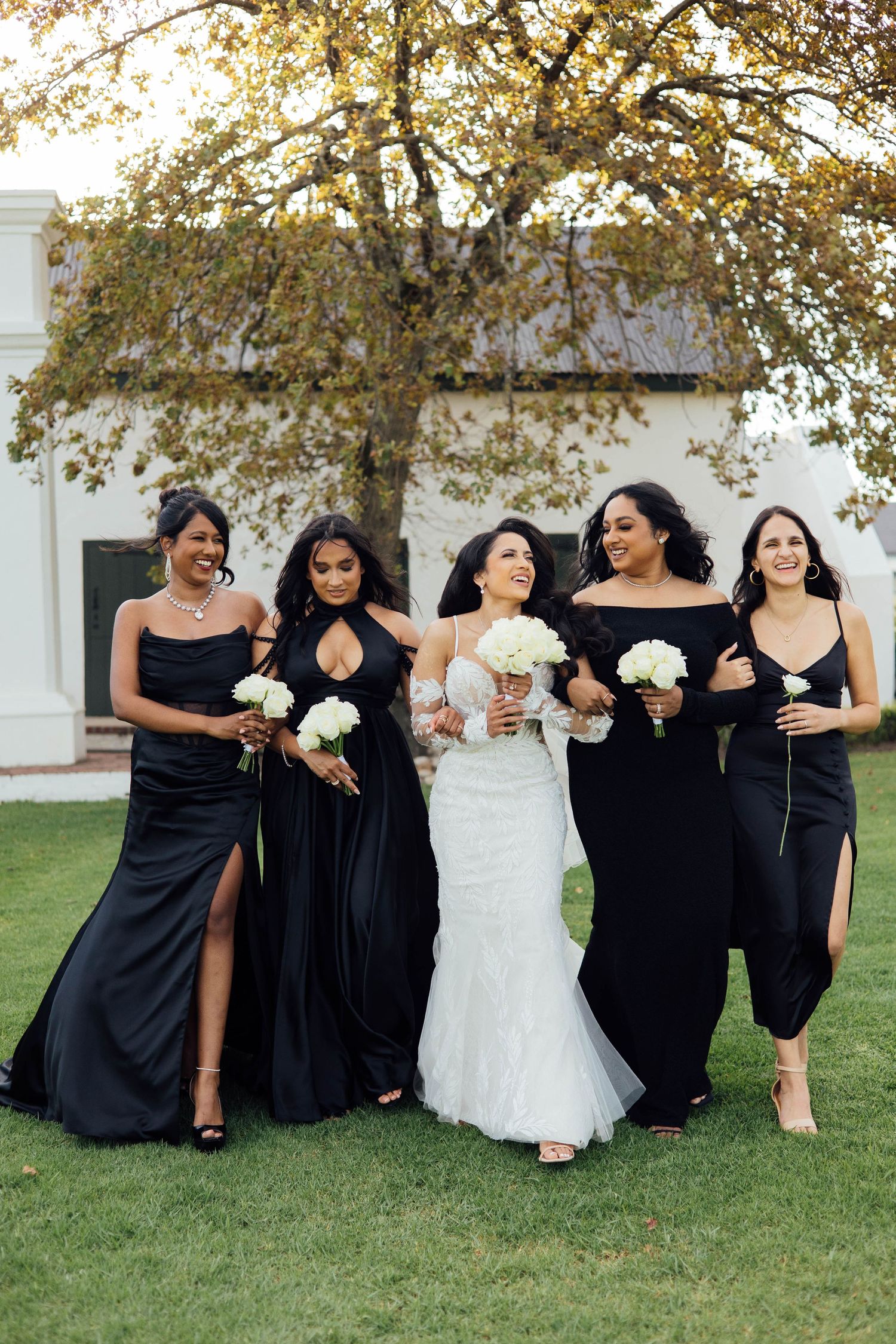 Wedding party wearing elegant black bridesmaid dresses and white bridal gown walk together on grass with tree in background.