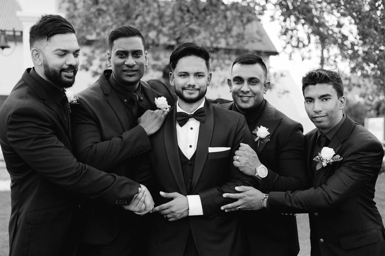 Black and white wedding photo showing a groom and groomsmen in formal attire laughing together outdoors.