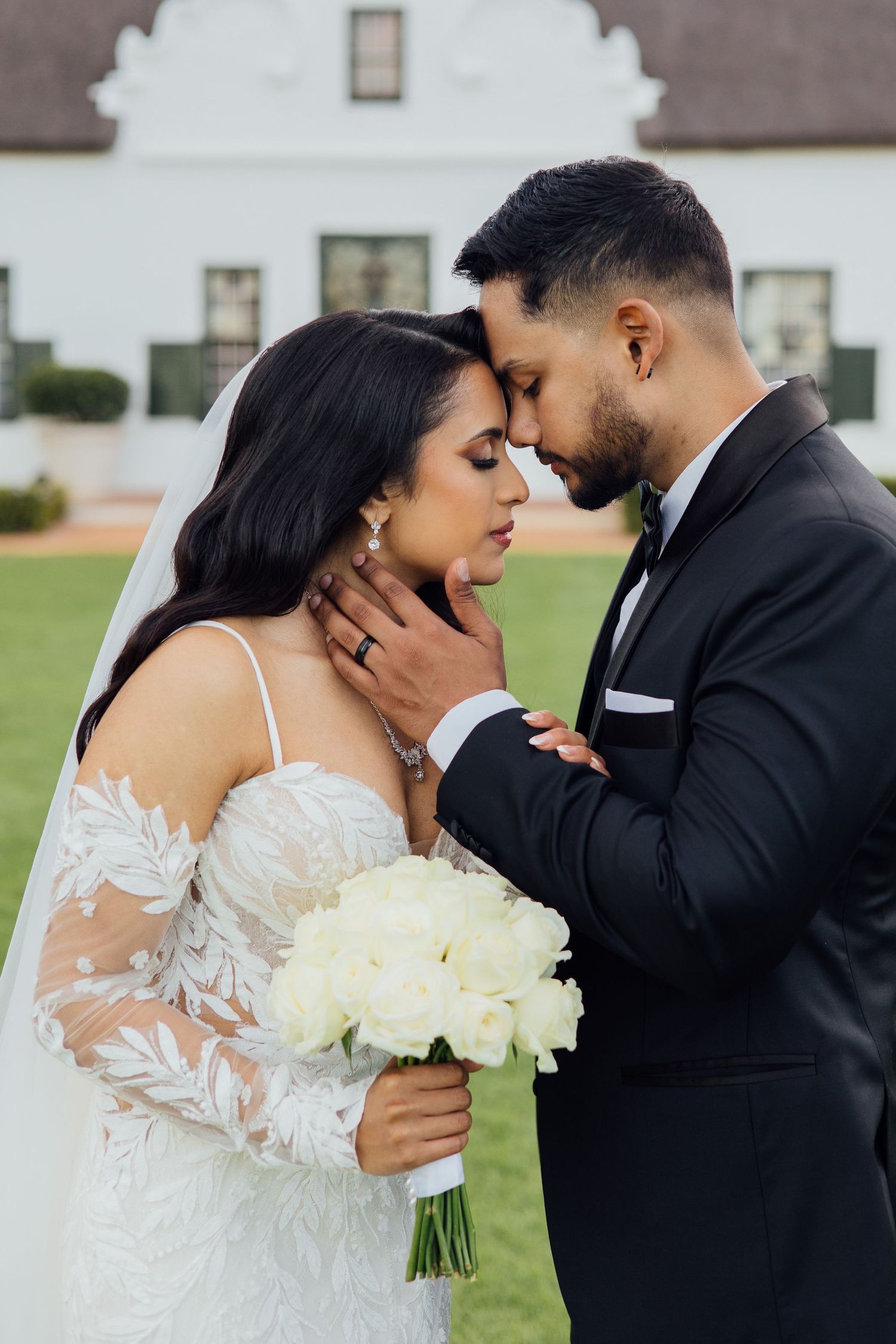 Romantic wedding couple embraces outdoors with white bouquet in front of historic white building.
