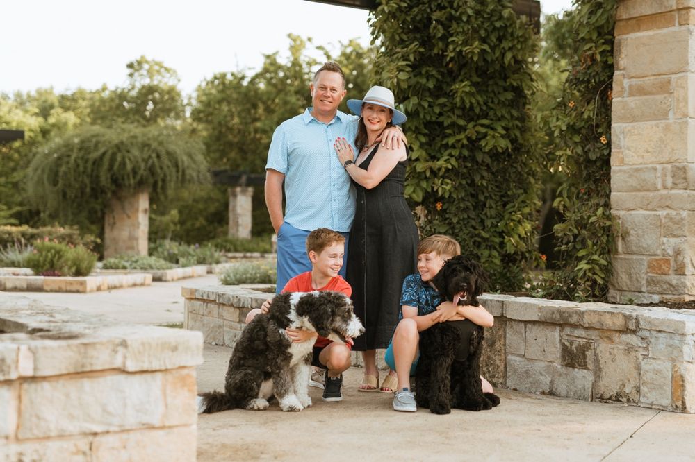 A family stands on stone steps in a garden setting with their two dogs on a sunny afternoon.